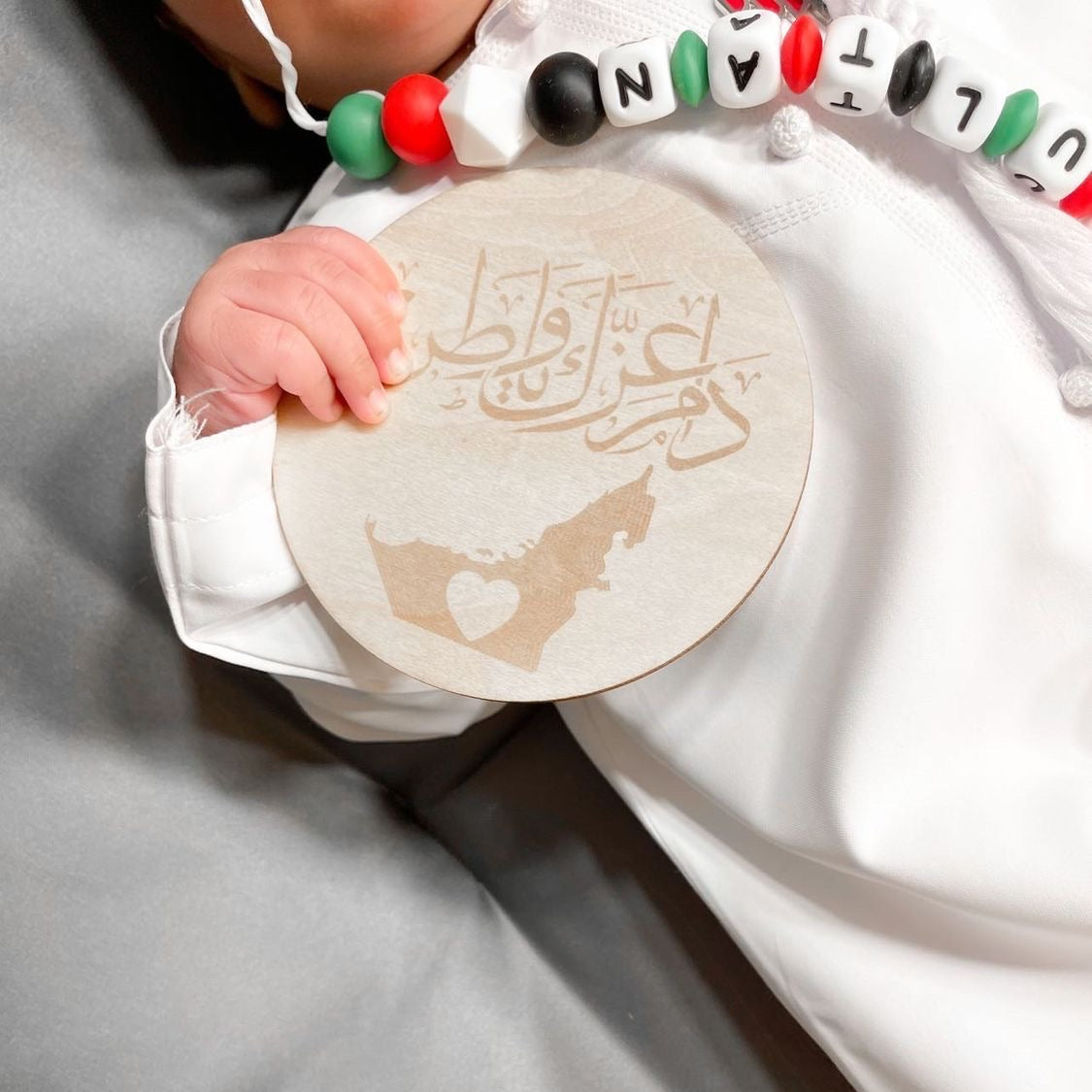 A close-up of a baby wearing a white traditional outfit and a colorful beaded necklace, holding a round wooden plaque engraved with Arabic calligraphy and a UAE map FOR UAE NATIONAL DAY IN DUBAI .