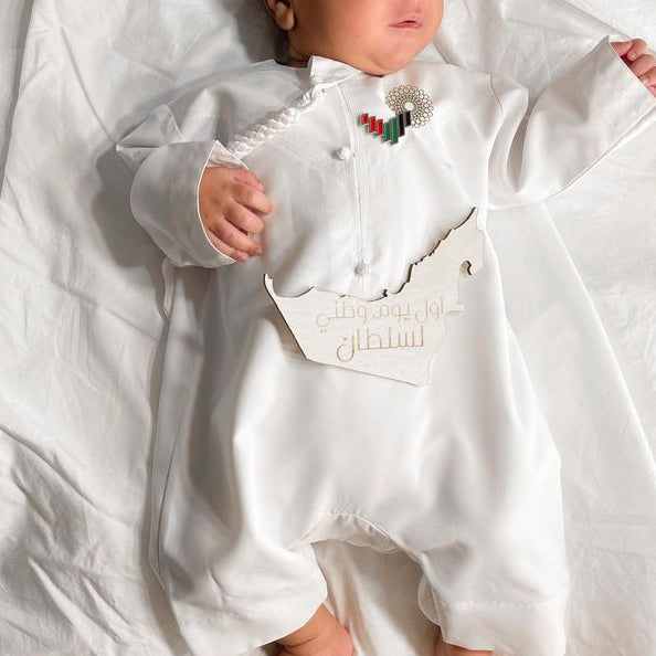 A baby lying on white bedding, wearing a white long-sleeve baby jumpsuit with decorative trim, a small colorful chest emblem, and a white bib with gold lettering.