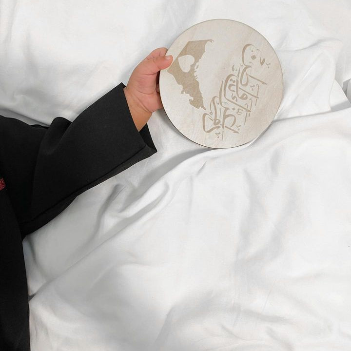 A hand holding a round ceramic plate with a beige map of EMIRATI and Arabic calligraphy, set against a background of white bedding.