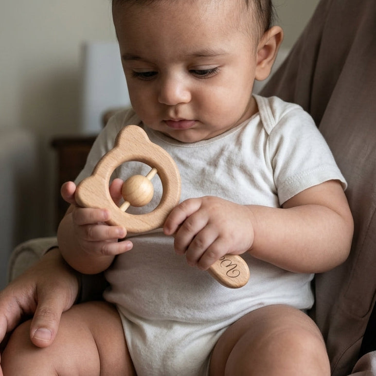 A baby sitting on an adult's lap holds a light natural wood bear-shaped teether toy with a ball in the center. The baby is wearing a white short-sleeve bodysuit in a softly lit indoor setting.