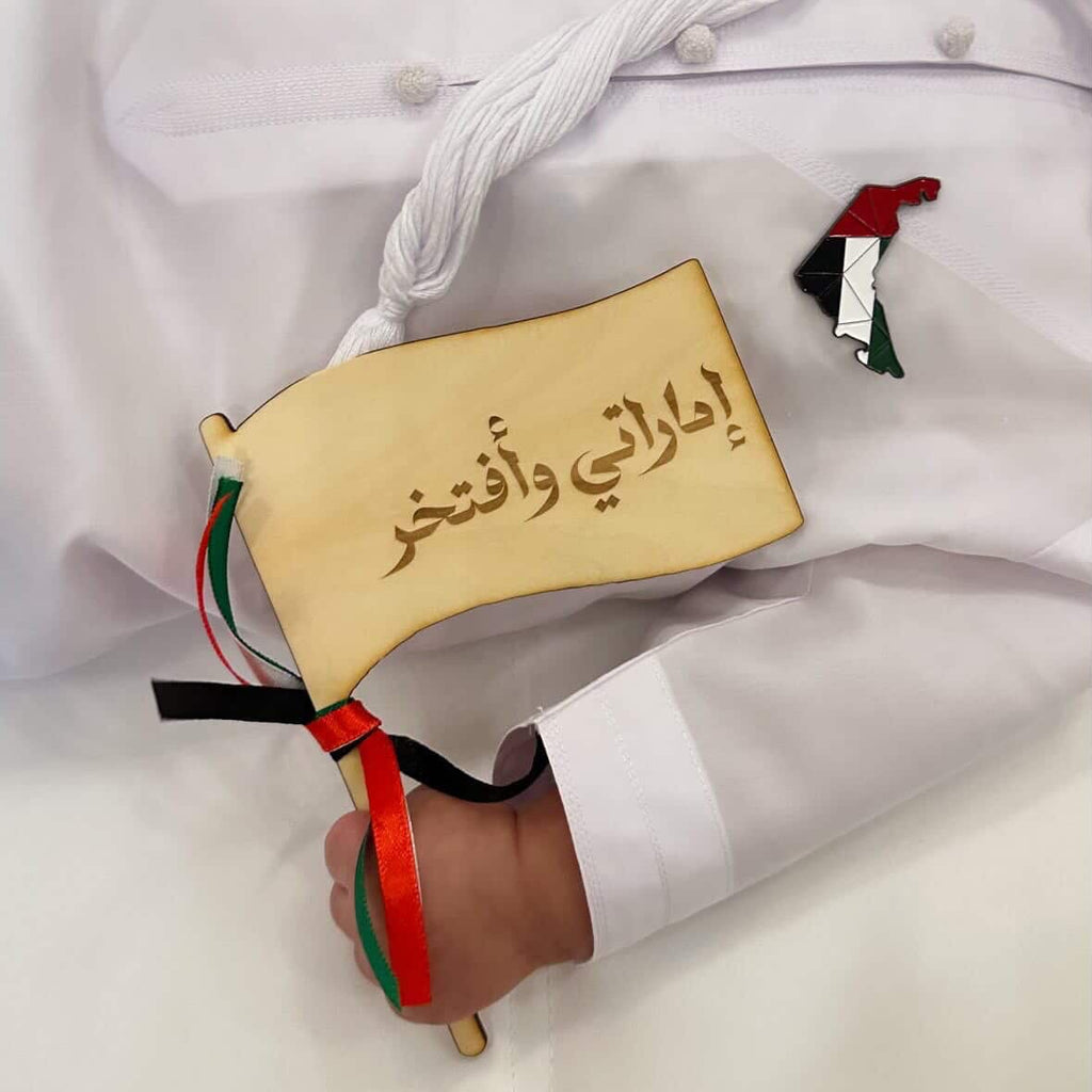 A baby wearing a white traditional thobe lies on a white surface, holding a small beige flag with Arabic calligraphy and red, green, and black ribbons. A small UAE flag pin is pinned to the chest.