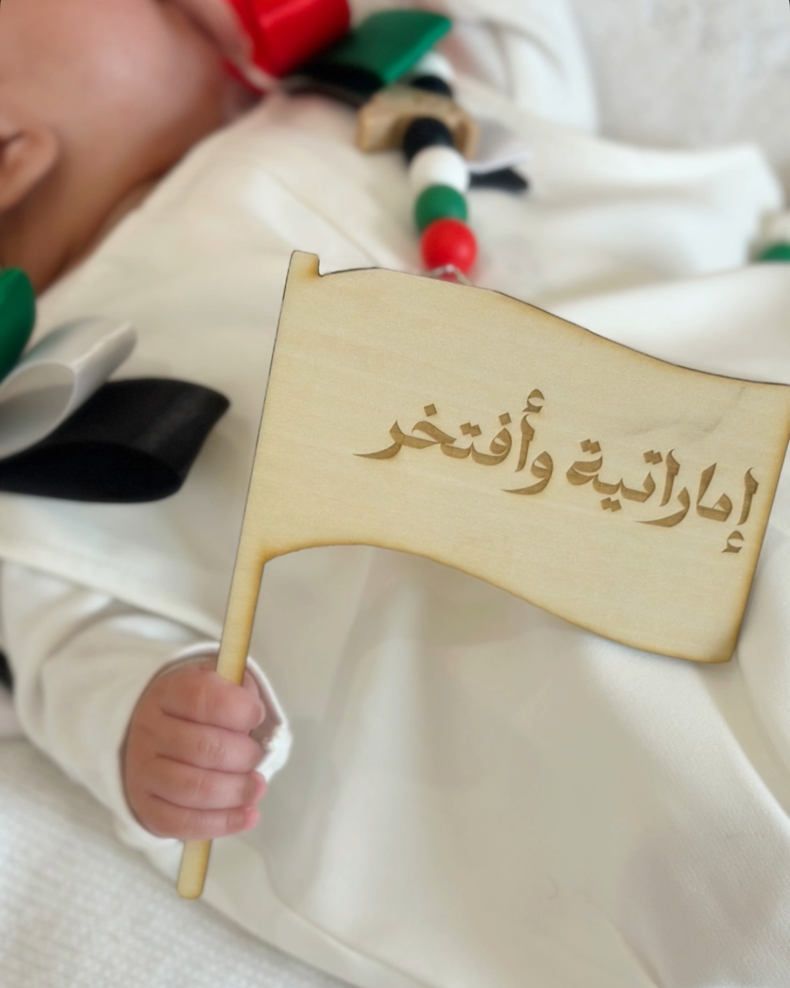 A close-up lifestyle shot of a baby wearing a white traditional outfit and a red, white, and green headband, holding a small wooden flag engraved with Arabic text.