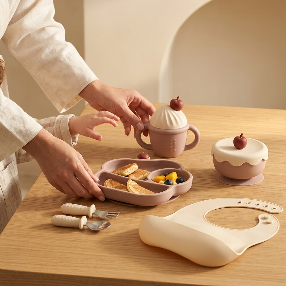 A styled tabletop scene showing a baby feeding set in warm neutral tones. A person in a cream linen outfit is placing a pastel pink divided plate with small food pieces into a small pink handled cup decorated like a dessert with a cherry on top.