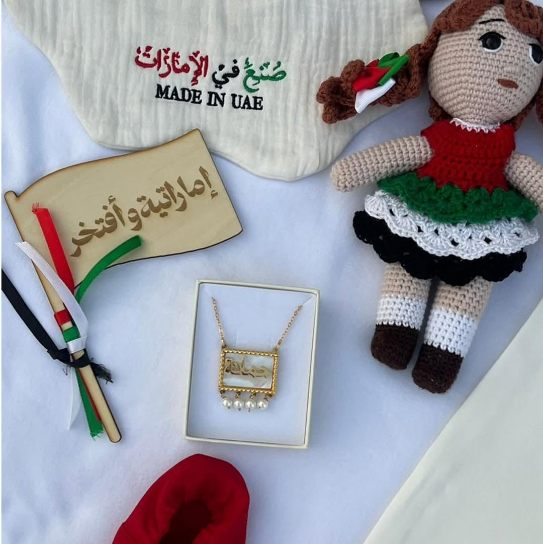 A baby gift set arranged on a white fabric background, including a cream-colored bonnet with 'MADE IN UAE' text, a crocheted doll in a red and green dress, a small gold necklace in a box, a wooden sign with Arabic text, red slippers, and a small flag.