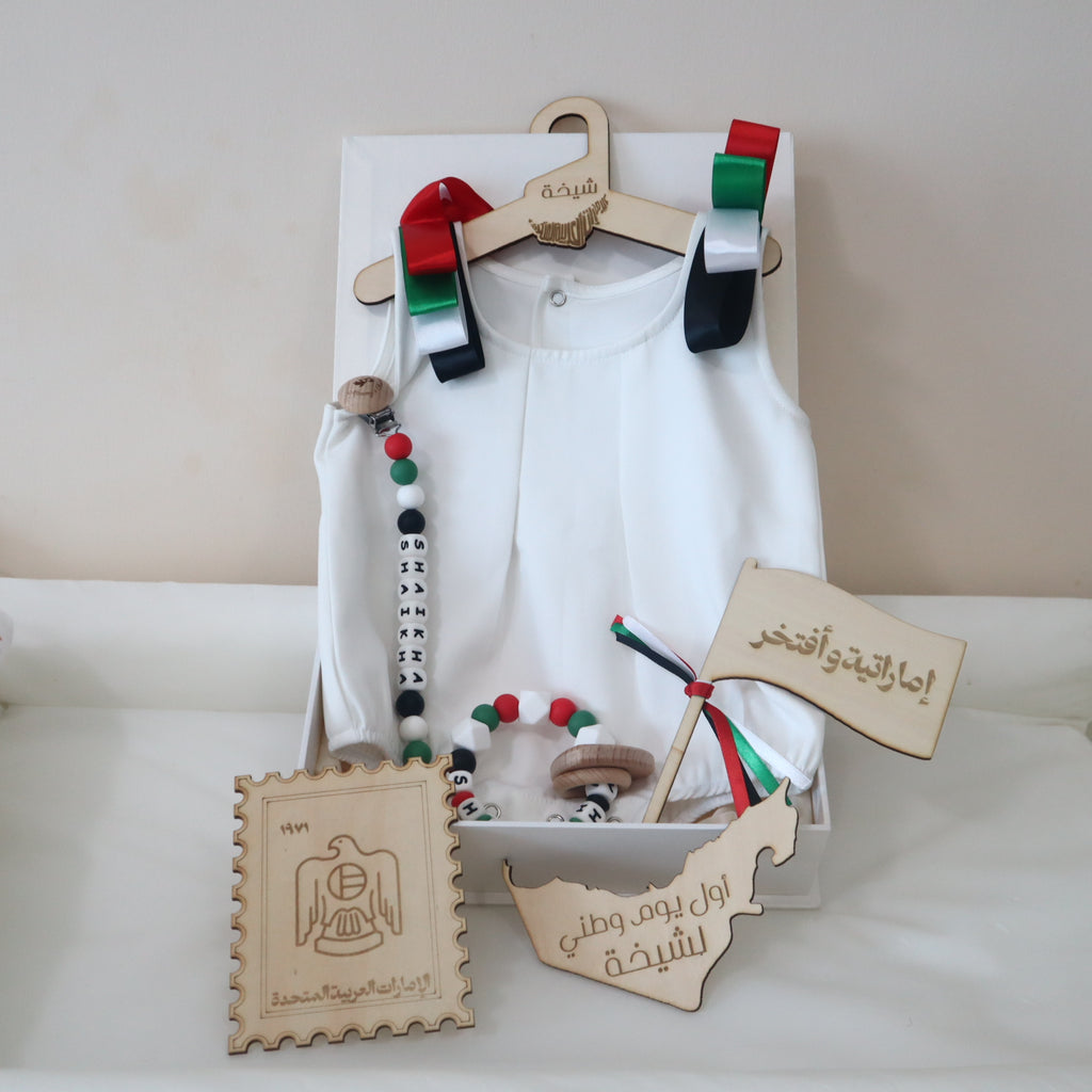 A baby boy's traditional Islamic outfit set displayed on a wooden hanger inside a white gift box. The set includes a white long-sleeve top, a white belly cloth, a wooden bead necklace, a wooden bead bracelet, a miniature Iraqi flag, a wooden stamp-shaped plaque, and two wooden map-shaped cards with Arabic calligraphy.