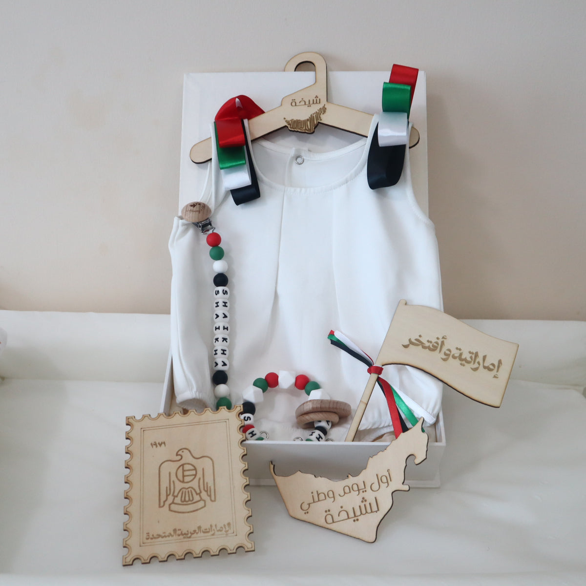 A baby boy's traditional Islamic outfit set displayed on a wooden hanger inside a white gift box. The set includes a white long-sleeve top, a white belly cloth, a wooden bead necklace, a wooden bead bracelet, a miniature Iraqi flag, a wooden stamp-shaped plaque, and two wooden map-shaped cards with Arabic calligraphy.
